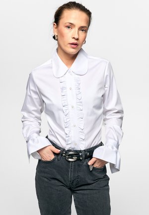Kvinne iført hvit skjorte med volanger og knapper, stor rundet krage, mansjetter på ermene, svart belte og mørke høytlivsjeans, hender i lommer.
