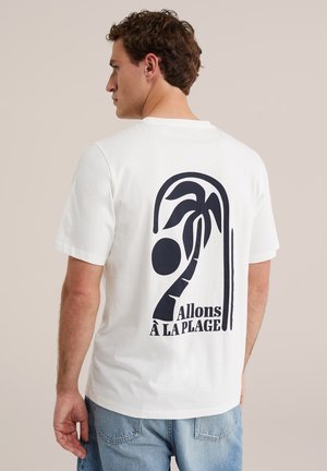Mann trägt weißes T-Shirt mit schwarzer Palmen-Grafik und dem Text „Allons à la plage“ auf dem Rücken, kombiniert mit blauen Jeans.