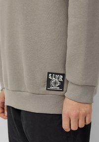 Beżowa bluza z rękawem i dołem w kolorze czarnym, z naszywką z napisem "SLVR Uwierz w swój potencjał na całym świecie", a ręka spoczywa obok czarnych spodni.