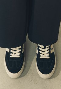 Mustat samettiset lenkkarit vaalean kermanvärisillä nauhoilla, joissa on retrotyylinen muotoilu, teksturoitu pinta ja kontrastiset sivuaksentit. Aseteltu vaalealle pinnalle.