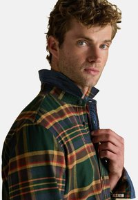 Vīrietis ar gaiši ziliem acīm un cirtainiem matiem pielāgo zaļo, sarkano un dzelteno raibiem krekliem ar džinsa iekšu apmali balta fona priekšā.