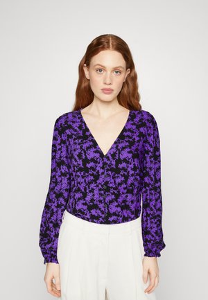 Une femme aux cheveux roux portant une blouse boutonnée à manches longues de couleur violette et noire à motifs floraux, associée à un pantalon blanc taille haute, faisant face à l'objectif.