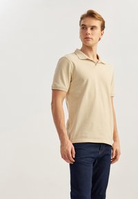 Polo beis con cuello clásico y mangas cortas, hecho de algodón. Combinado con jeans azul oscuro, de textura suave y diseño ajustado.