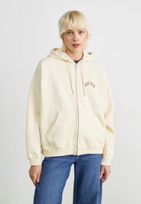 Kremfarget zip-hettegenser med frontlomme, snor i hetten og en liten brodert "Roxy"-logo. Mykt materiale og avslappet passform.