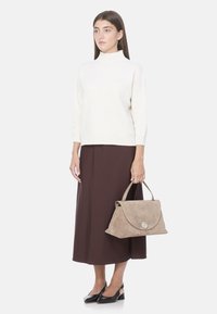 Borsa in suede beige con patta e hardware argentato, indossata da una persona con un maglione bianco e una gonna marrone, abbinata a scarpe nere.