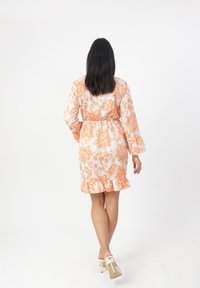 Korte mouwen jurk in een bloemenpatroon met oranje en witte kleuren, met een taille die is geaccentueerd en een gerimpelde zoom. Gekozen met lichtgekleurde hakken.