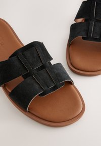 Sandales en daim noir à deux larges brides, présentant un design géométrique. La semelle est lisse et marron, avec une forme profilée pour le confort.