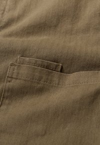 Braunes Fischgrät-Gewebe mit strukturiertem Erscheinungsbild, das auf der rechten Seite ein kleines Taschendetail aufweist und feine Musterlinien einfängt.