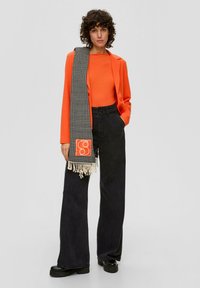 Frau mit lockigem Haar, die ein orangefarbenes Oberteil und eine Jacke, schwarze weit geschnittene Hosen, schwarze Schuhe und einen schwarz-weißen Hahnentritt-Schal mit orangefarbenem Patch trägt.