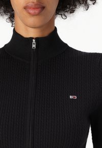 Černý pletený svetr na zip s vroubkovanou strukturou a vysokým límcem. Obsahuje malé vyšívané logo v červené, bílé a modré barvě na levé části hrudníku.