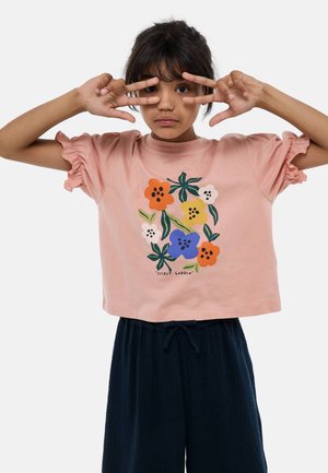 T-shirt rose court avec des manches volantes, ornée d'un motif floral en orange, bleu, jaune et vert. Inscription : "JARDIN SECRET."