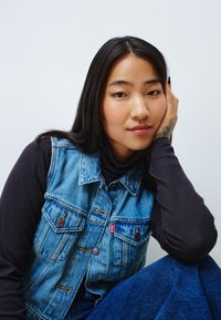 Gilet en denim avec une teinte bleu, présentant de grandes poches et des boutons marron, superposé sur un col roulé noir. Le mannequin a de longs cheveux foncés.