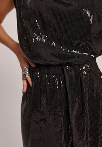 Zbliżenie na osobę w czarnej sukience ozdobionej cekinami, z wysmukloną talią, ręką opartą na biodrze i dużym srebrnym pierścionkiem na palcu.