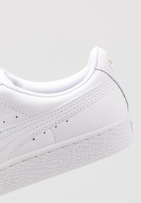 Bílé kožené tenisky s texturovanou gumovou podrážkou, perforovanými detaily a kontrastními šedými akcenty v oblasti paty.