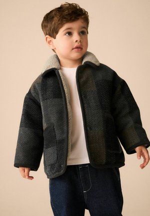 Junger Junge mit kurzen braunen Haaren, der eine dunkel karierte Jacke mit Fleece-Kragen über einem weißen Hemd und dunklen Jeans trägt, schaut zur Seite.