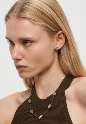 Blonde Frau trägt herzförmige goldene Ohrringe und eine passende goldene Halskette mit transparenten herzförmigen Anhängern, in einem braunen ärmellosen Oberteil.