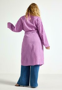 Lang lilla skjorte med lodrette striber, knapmanchetter og indsnørret talje. Kombineret med vide blå jeans og flade sandaler.