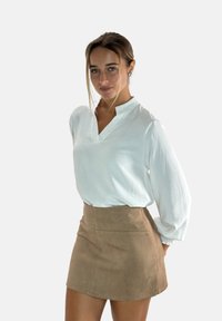 Weiße, strukturierte Bluse mit einem tiefen Kragen, kombiniert mit einem beigen Wildleder-Minirock, der eine figurschmeichelnde Silhouette und seitliche Taschen hat.