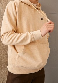 Beige hoodie with speckled texture, adjustable drawstring, kangaroo pocket, and black Nike logo on the chest, paired with dark pants.