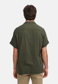 Chemise à manches courtes en tissu texturé vert foncé, avec un col et un ourlet droit. Le dos présente un motif de rayures verticales. Pas de matériel visible.