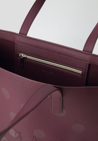Bordeaux tas van textuurmateriaal, met een ritsvak aan de binnenkant en een reliëflogo, accentueert grote, subtiele stippen.