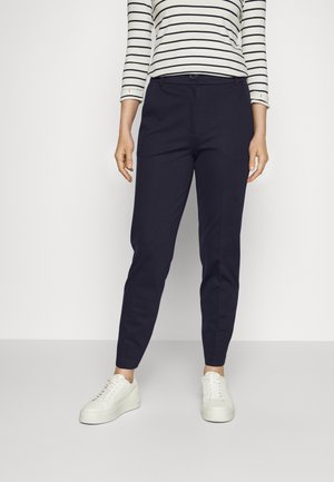 Femme portant un pantalon slim bleu marine, des baskets compensées blanches et une chemise blanche à manches longues avec des rayures horizontales noires.