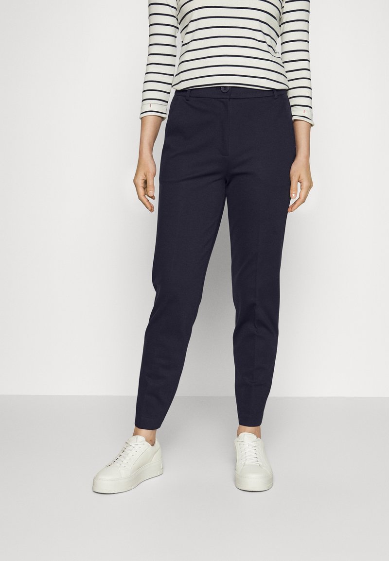 Femme portant un pantalon slim bleu marine, des baskets compensées blanches et une chemise blanche à manches longues avec des rayures horizontales noires.