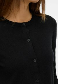 Femme portant un cardigan noir à boutons avec un col rond, montrée de la nuque jusqu'au milieu du torse sur un fond uni.