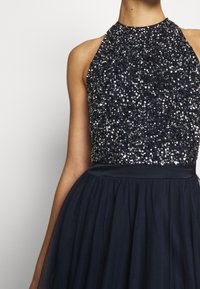 Lauluva dressi, jossa on helmikirjailtu, olkaimeton halterneck-paita ja virtaava, teksturoitu hame. Kimaltavat koristeet yksityiskohtaisena yläosassa.