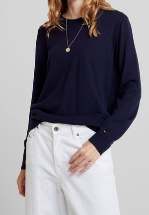 Kvinde iført en navyblå langærmet sweater, hvide bukser og en guldkæde, med hånden hvilende på hoften.