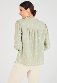 Blusa verde clara hecha de un tejido suave, con cuello con volantes, mangas abullonadas y patrones de hojas blancas en toda la prenda.