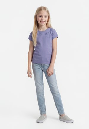 Smilende jente med langt blond hår iført en lilla kortermet skjorte, lyseblå jeans og grå slip-on sko, stående i en avslappet positur.