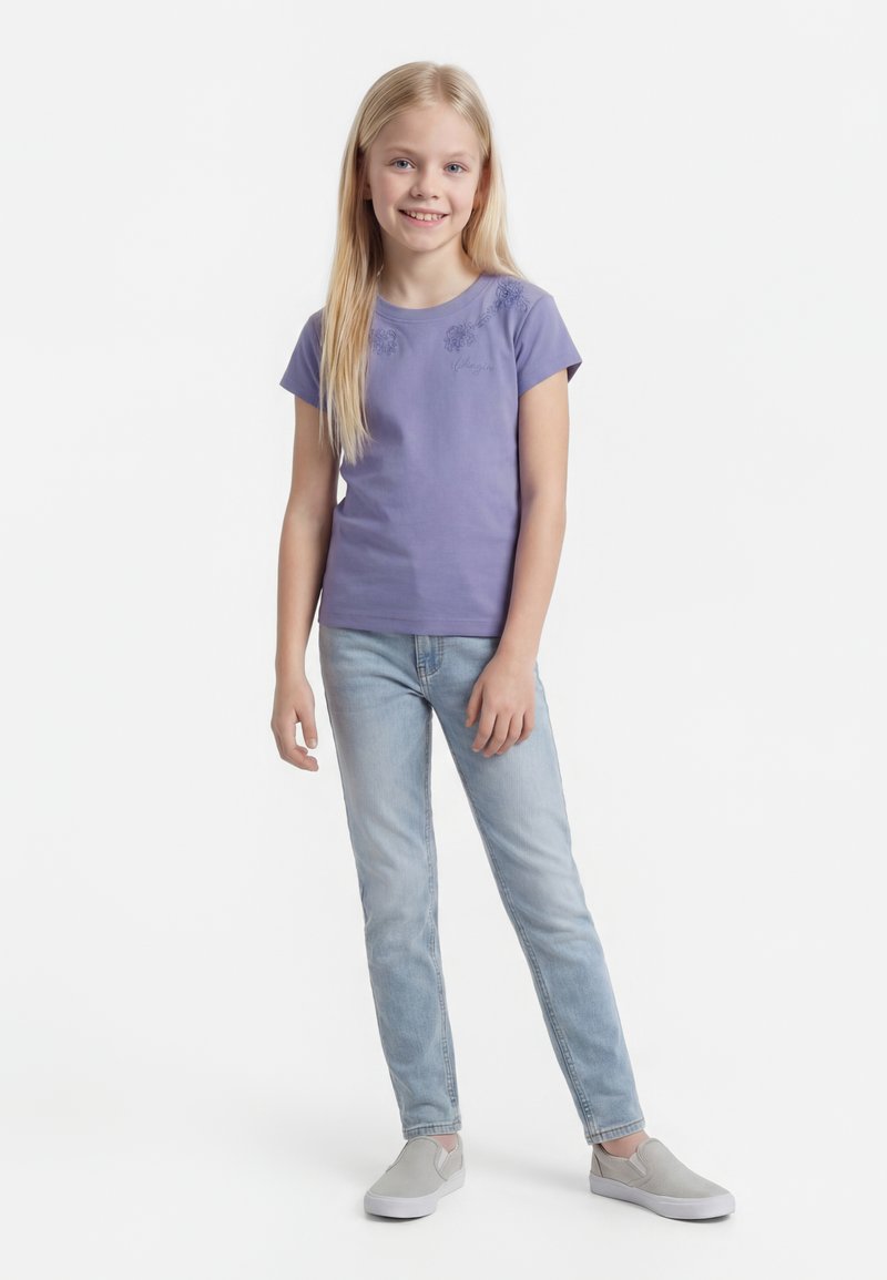 Fille souriante aux longs cheveux blonds vêtue d'un t-shirt violet à manches courtes, d'un jean bleu clair et de chaussures grises sans lacets, debout dans une pose détendue.