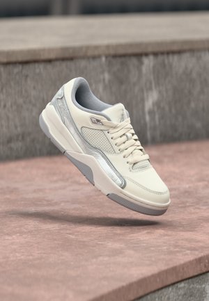 Valge ja halli värvi sportlik toss, millel on paeltega kinnitamine, pehmendatud krae ja tekstureeritud tald, kujutatud hõljumas betoonpinna kohal.