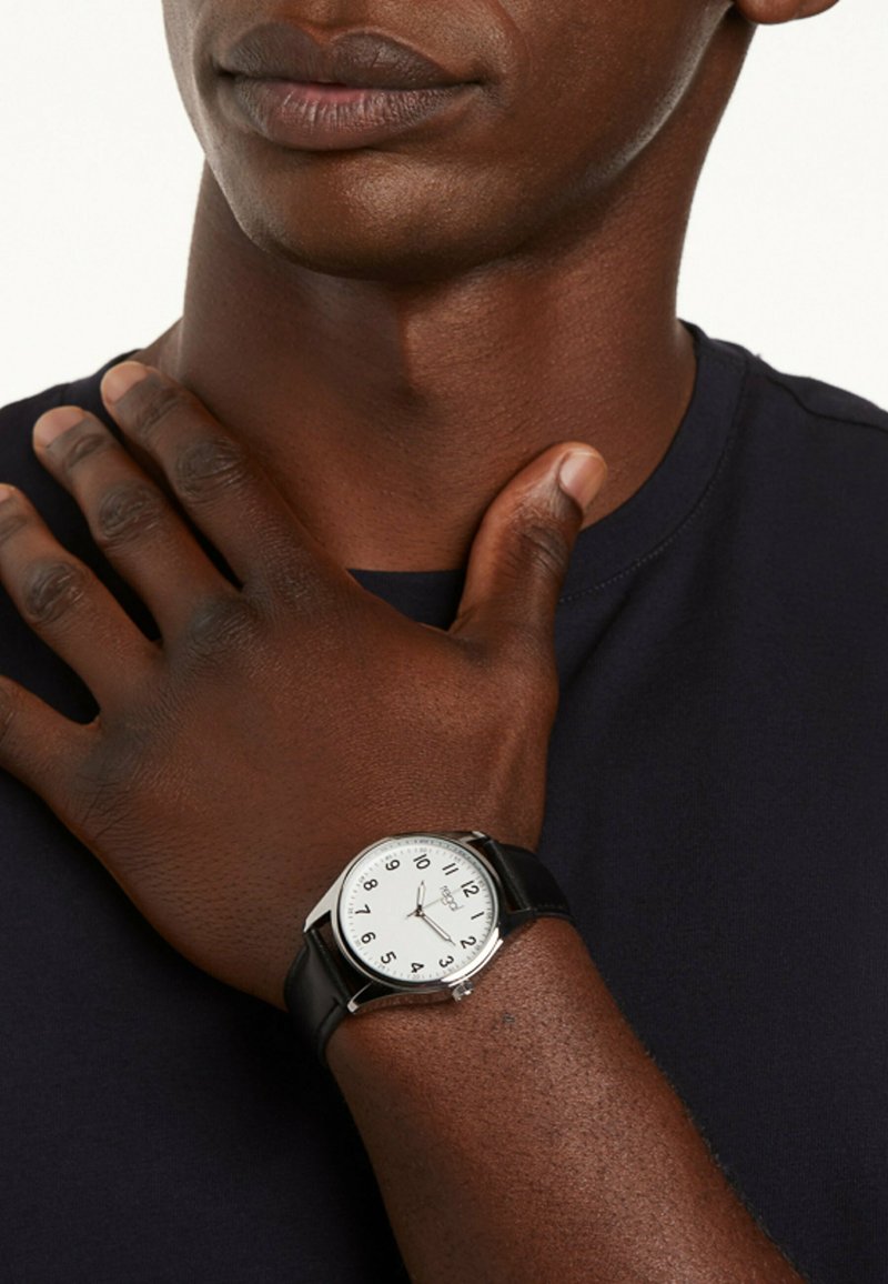 Round wristwatch with a stainless steel case, white dial, black numbers, and a black leather strap. Visible hand resting on neckline.