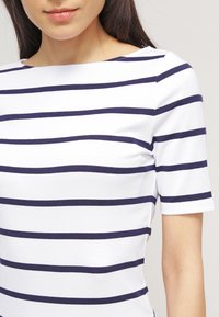 Vit topp med mörkblå horisontella ränder, rund halsringning, korta ärmar och en figursydd design, tillverkad av ett stretchigt tyg.