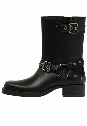 Botín de cuero negro hasta el tobillo con punta redonda, que presenta correas ajustables con hebillas metálicas y tachuelas, y un tacón bajo tipo bloque.