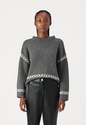 Grå strikket genser med rund hals og kontrasterende hvite broderimønstre på ermene og nederkanten, kombinert med svarte bukser.