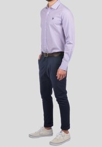 Man wearing light purple checkered button-up shirt, dark blue trousers with brown belt, and beige sneakers, standing against a plain background.