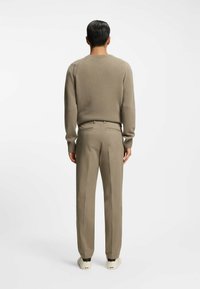 Beige Wollpullover mit enganliegenden Ärmelbündchen, kombiniert mit maßgeschneiderter beige Hose. Die Schuhe sind cremefarben mit schwarzen Akzenten. Sauberes und einfaches Design.