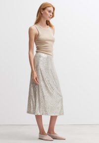 Beige ärmelloses Oberteil, kombiniert mit einem knielangen silbernen Paillettenrock mit vertikalem Linienmuster und transparenten flachen Schuhen.