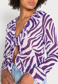 Blusa com estampa de zebra em roxo e branco, apresentando detalhes em babados, um decote em V profundo e mangas longas. O material é semi-translúcido com uma textura leve.
