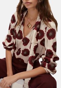 Blusa di seta con scollatura a V profonda, caratterizzata da un motivo floreale bordeaux su uno sfondo crema. Abbinata a pantaloni borgogna scuri e collana dorata.