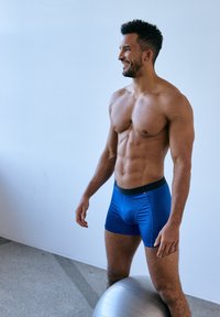 Blå boxerkalsonger med en svart midjeresår som har varumärkestryck, tillverkade av ett mjukt tyg. Modellen står bredvid en silverfärgad träningsboll.