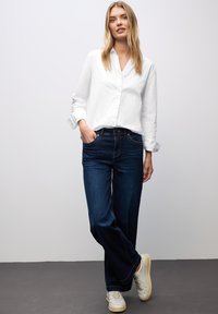 Camisa blanca de manga larga con botones, combinada con vaqueros de pierna ancha de color azul oscuro. Zapatillas blancas con suelas claras. Texturas suaves, ajuste informal.