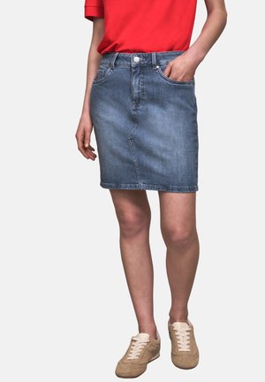 Personne portant une jupe en denim bleu jusqu'aux genoux, une chemise rouge, des baskets beige, une main dans la poche de la jupe, debout devant un fond blanc.