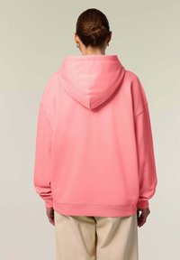 Felpa oversize rosa con cappuccio regolabile, polsini a costine e una texture liscia, abbinata a pantaloni beige, vista di spalle.