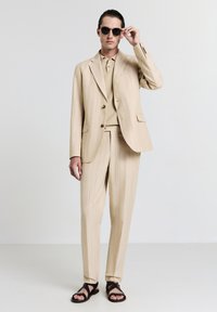 Beige gestreifter Anzug mit einem einreihigen Blazer, passenden Hosen und einem strukturierten Poloshirt darunter. Kombiniert mit schwarzen runden Sonnenbrillen und Sandalen.