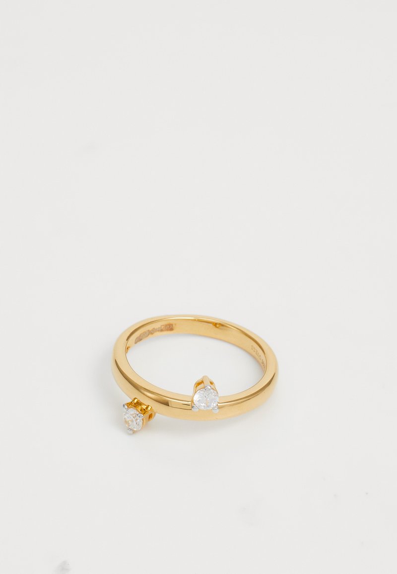 Anel de metal dourado com duas pedras preciosas redondas e transparentes fixadas em configurações de garras. Banda circular lisa com acabamento polido e textura subtile.
