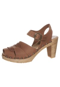 Sandale en cuir marron avec un talon épais, bout ouvert et sangle à la cheville. Présente des lanières croisées et des clous en métal le long de la semelle. Base en bois texturé.